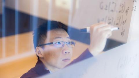 Georgia Tech Assistant Professor Glen Chou with the School of Cybersecurity and Privacy works through an equation on a transparent writing board.